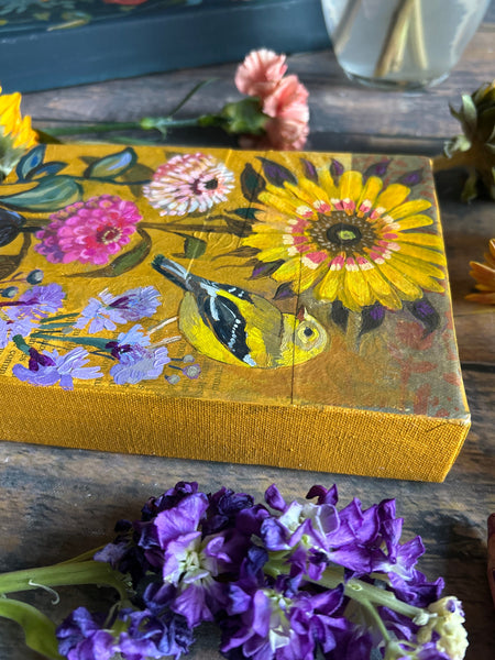 Decorative notebook with floral and bird design on a wooden surface with flowers around